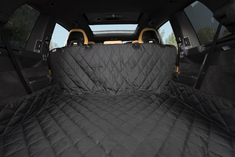 Car trunk lined with a black, quilted cover, viewed from the backseat. The interior appears spacious with visible rear seat backs. Sunlit trees are seen through the windows.
